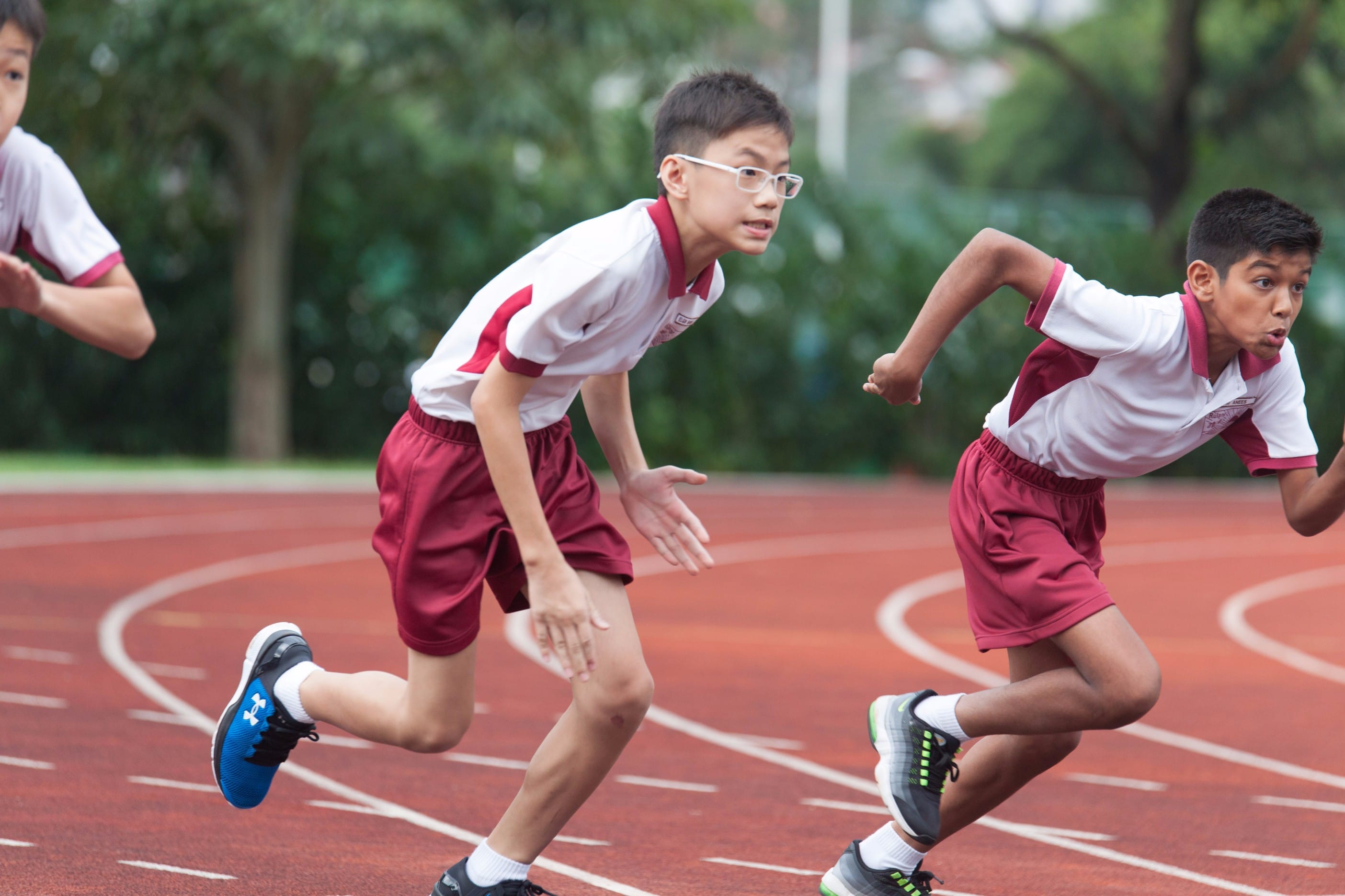 Three boys in matching white and red uniforms sprint on a red running track surrounded by green trees.